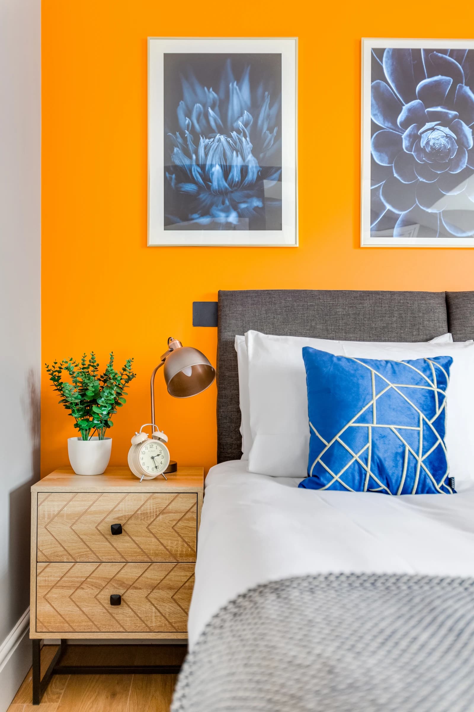 Bedside table with plant, lamp and alarm clock against orange wall at Boon House, Egham