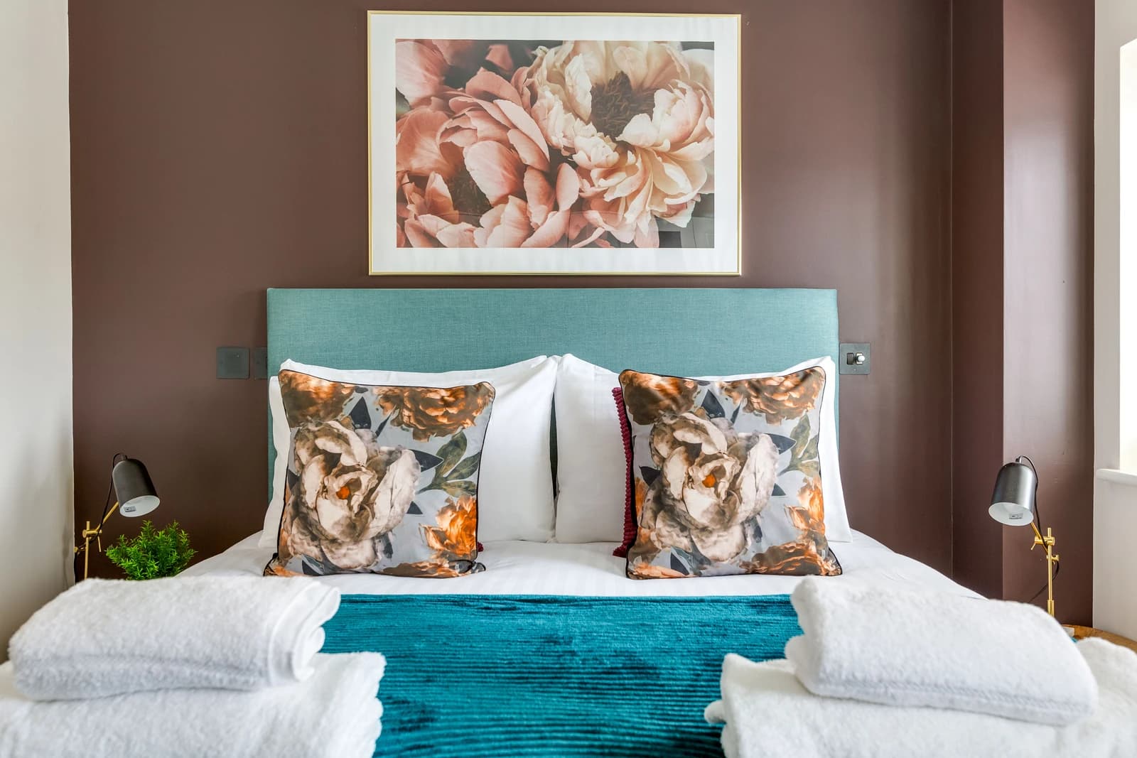 King-size bed with teal throw, floral cushions and artwork at Boon House serviced apartment, Egham